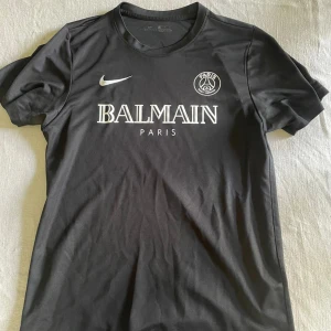 Svart PSG fotbollströja Nike x Balmain - Säljer en svart fotbollströja från Nike i samarbete med PSG och Balmain. Tröjan har korta ärmar, rund hals och tryck med Balmain Paris samt PSG-logga på bröstet. Materialet är lätt och andas, perfekt för fotboll eller träning.