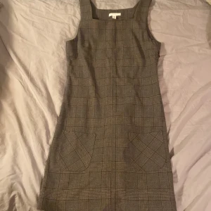 Rutig ärmlös klänning i ullblandning - Snygg ärmlös klänning med rutigt mönster i grå och bruna toner. Klänningen har fyrkantig ringning och två fickor framtill. Materialet känns som ullblandning och modellen är figurnära upptill med lätt utställd kjol.