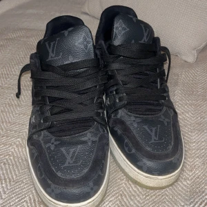Svarta Louis Vuitton sneakers med monogram - Säljer ett par svarta Louis Vuitton sneakers med klassiskt monogrammönster över hela skon. Skorna har svarta snören, vit sula och tydliga LV-detaljer. Tillverkade i Italien och har en modern, sportig look.