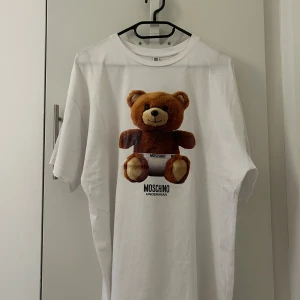 Vit t-shirt med nalletryck från Moschino - Vit t-shirt från Moschino Underwear med ett stort tryck av en söt brun nalle på framsidan. Klassisk passform och rund halsringning. Tillverkad i 100% bomull för en mjuk och skön känsla. Perfekt statement-plagg för dig som gillar lekfull stil.