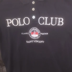 Svart pikétröja från Polo Club Saint Vincent - Klassisk svart pikétröja från Polo Club Saint Vincent med broderad logga och text på bröstet. Tröjan har tre knappar vid halsen och korta ärmar. Perfekt för en stilren och sportig look.