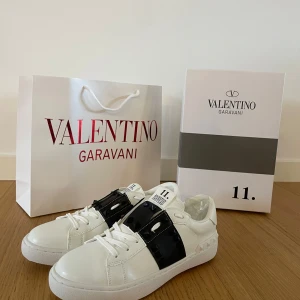 Valentino Garavani Rockstud Untitled sneakers - Säljer ett par vita Valentino Garavani Rockstud Untitled sneakers med svart band och ikoniska nitar över vristen. Skorna har rund tå, platt sula och snörning. Märkeslogga på plösen och innersulan. Riktigt snygga och trendiga sneakers!