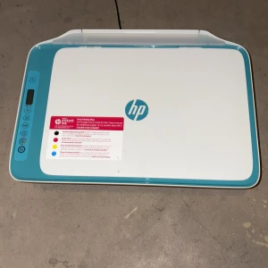 HP multifunktionsskrivare med skanner - Säljer en vit och turkos HP multifunktionsskrivare med inbyggd skanner. Skrivaren har en digital display och flera funktionsknappar på vänster sida. Passar för både utskrift och scanning av dokument. HP-logga synlig på ovansidan.