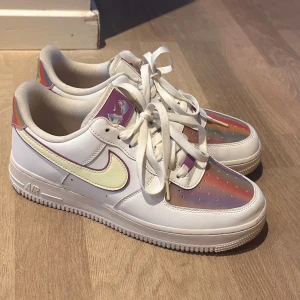 Nike Air Force 1 med regnbågsskimrande detaljer - Säljer ett par vita Nike Air Force 1 med unika regnbågsskimrande paneler framtill och på hälen. Skorna har klassisk snörning, rund tå och en vit sula. Nike loggan  är i ljusgul nyans och materialet är syntet och skinn.