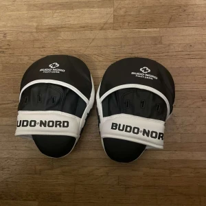  träningsmittsar Budo-Nord - Säljer ett par svarta och vita träningsmittsar från Budo-Nord. Perfekta för kampsportsträning med en bekväm passform och robust design. Mittsarna är justerbara remmar för säker användning.