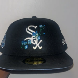 Svart keps från New Era x White Sox med blommor - Svart keps med White Sox-logga framtill, dekorerad med blå broderade blommor. Kepsen har platt skärm, broderade detaljer och patch på sidan med 'World Series'. MLB-logga baktill. Perfekt för dig som gillar sportig stil med unik twist.