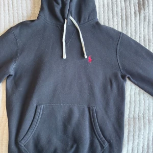 Svart hoodie från Polo Ralph Lauren - Svart hoodie från Polo Ralph Lauren med klassisk huva och vit dragsko. Tröjan har en röd broderad logga på bröstet och en stor magficka framtill. Perfekt för en avslappnad stil. Kontakta mig vid frågor🙋‍♂️9.5/10 skick!