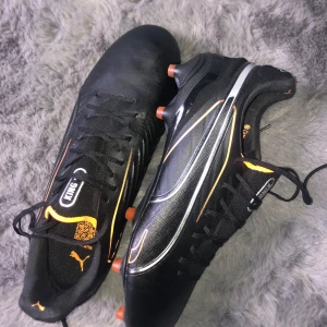 Svart  elite fotbollsskor från Puma med orange detaljer -   ⚽️ Grymma fotbollsskor från Puma säljes! ⚽️  Snygga svarta Puma-skor med orange detaljer och logga. Perfekt för dig som vill sticka ut på planen! Skorna har: - Snörning - Platt sula med dobbar för bra grepp - Väska medföljer  Använda en gång, så de är i toppskick. Perfekt om du vill ha ett par riktigt bra skor till ett bra pris!