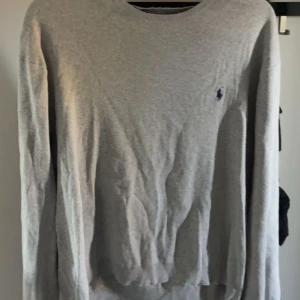 Ljusgrå tröja från Ralph Lauren - Säljer en ljusgrå stickad tröja från Ralph Lauren med rund halsringning och klassisk broderad logga på bröstet. Tröjan har lång ärm och är perfekt för lager-på-lager-stil. Enkel och stilren design som passar till mycket.