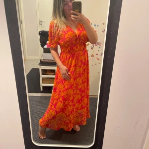Blommig långklänning i orange och rosa - Säljer en färgstark långklänning med blommigt mönster i orange och rosa. Klänningen har v-ringning, korta ärmar och markerad midja. Perfekt för dig som vill sticka ut med en somrig look!Storlek pasar både M och L 38 och 40 halt ny