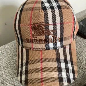 Beige rutig keps från Burberry - Säljer en klassisk keps från Burberry i beige med det ikoniska rutmönstret och broderad logga framtill. Kepsen har böjd skärm och justerbar passform. Perfekt accessoar för en stilren look.