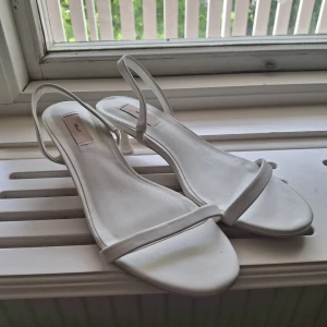 Vita låga klackar från Nelly - Säljer ett par stilrena vita klackar från Nelly med smal rem över tårna och runt hälen. Klacken är låg. Passar perfekt till bal, skolavslutning eller student! Använd ett fåtal gånger. 💞