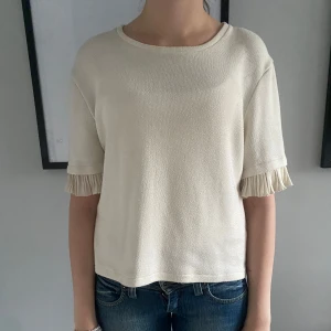 Beige topp med volangärmar - Säljer en beige topp från zara med rund halsringning och korta ärmar med volangdetalj. Toppen har en rak passform och är tillverkad i ett mjukt material som känns skönt mot huden. Perfekt för en stilren och enkel look. Tröjan finns inte längre att köpa.