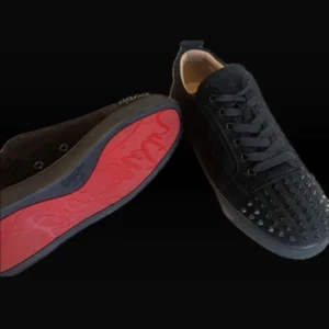 Christian Loubitins - Säljer ett par svarta sneakers i mocka från Christian Louboutin med ikonisk röd sula och svarta nitar på tån. Skorna har snörning och en platt sula. Perfekta för dig som vill sticka ut med en lyxig och edgy stil.