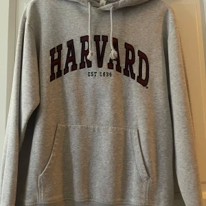 Grå Harvard hoodie - Säljer en grå hoodie med stor HARVARD-logga i vinrött och svart på bröstet. Tröjan har huva med dragsko och en stor magficka. Perfekt för dig som gillar college-stil!