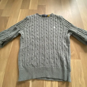Grå kabelstickad tröja från Polo Ralph Lauren - Säljer en klassisk grå kabelstickad tröja från Polo Ralph Lauren med rund halsringning och broderad logga på bröstet. Tröjan har långa ärmar och breda ribbade muddar vid ärmslut och nederkant. Perfekt för kyligare dagar. Står att det är strl l men passar ungefär 168-175 cm. Får även med en polo plast påse 