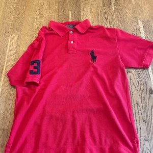 Röd pikétröja från Polo Ralph Lauren - Klassisk röd pikétröja från Polo Ralph Lauren med korta ärmar och krage. Broderad logga på bröstet och siffra 3 på ärmen. Perfekt för en sportig och stilren look.