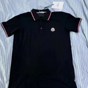 Svart pikétröja från Moncler - Säljer en svart pikétröja från Moncler med klassisk krage och knäppning framtill. Tröjan har Moncler-logga på bröstet och röda, vita och blå detaljer på krage och ärmslut. Perfekt för en stilren och sportig look.