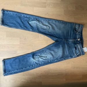 Blå Replay jeans straight fit - Säljer ett par klassiska blå jeans från Replay med raka ben och snygga slitningar. Jeansen har fem fickor, dragkedja och knapp i midjan samt Replay-logga på fickan. Perfekta för en avslappnad stil.
