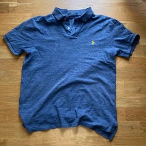 Blå pikétröja från Polo Ralph Lauren - Storlek L Slim som passar M! Säljer en blå pikétröja från Polo Ralph Lauren i slim fit-modell. Tröjan har korta ärmar, klassisk krage och knäppning framtill. Den är dekorerad med det ikoniska gula broderade logotypen på bröstet.