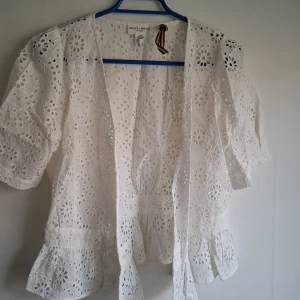 Vit broderad blus från Holly White - Säljer en söt vit blus från Holly White med broderade blommor och volangkant nedtill. Blusen har korta ärmar och är öppen framtill, perfekt för lager-på-lager. Superfin till sommaren!