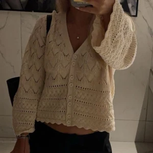 Beige stickad kofta med spetsdetaljer - Söt beige stickad kofta med v-ringning och knappar framtill. Koftan har långa ärmar och ett spetsliknande mönster samt vågig nederkant. Perfekt för lager-på-lager och en mysig look.