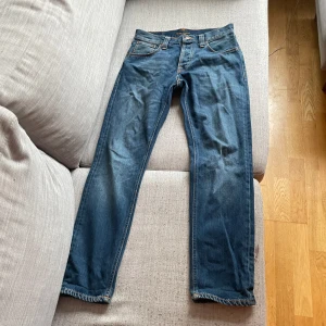 Blå jeans från Nudie Jeans Co - Säljer ett par klassiska blå jeans från Nudie Jeans Co med raka ben och snygga kontrastsömmar. Jeansen har fem fickor, knappgylf och läderpatch bak i midjan. Perfekta för dig som gillar stilrena och tidlösa jeans.