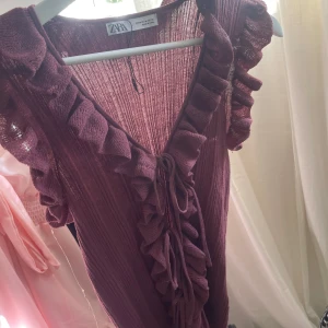 Vinröd volangblus från Zara - Supersöt vinröd blus från Zara med volangdetaljer längs ärmar och v-ringning. Blusen har knyt framtill och är tillverkad i ett ribbat material som ger en snygg struktur. Perfekt för dig som vill sticka ut med en romantisk look.