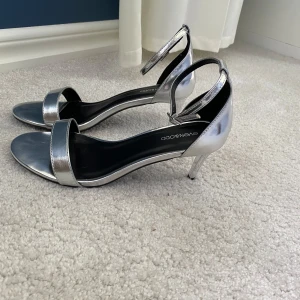Silvriga klackskor - Säljer ett par stilrena silverfärgade klackskor från Even&Odd. Skorna har öppen tå, smal rem över foten och justerbar ankelrem. Perfekta för att lyfta din outfit med en glansig touch.
