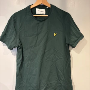 Grön t-shirt från Lyle & Scott - Säljer en mörkgrön t-shirt från Lyle & Scott med klassisk rund hals och korta ärmar. T-shirten har en broderad gul logotyp på bröstet och är tillverkad i mjuk bomull. Perfekt för en avslappnad stil.