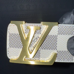 Louis Vuitton Damier-bälte med guldspänne - Säljer ett Louis Vuitton-bälte med klassiskt Damier-mönster i vitt och grått. Bältet har ett stort LV-spänne i guld och är tillverkat i skinn med beige insida. Perfekt accessoar för att lyfta din outfit.
