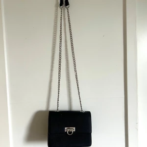 Svart axelväska med kedja - Snygg svart axelväska med silvrig kedja. En klassisk detalj som kan lyfta liken outfit som helst. Två fack + dragkedja inuti för kort eller liknande.
