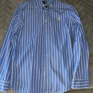 Blåvit randig skjorta från Polo Ralph Lauren - Snygg blå skjorta med vita ränder från Polo Ralph Lauren. Slim fit-modell med klassisk krage och knappar framtill. Ikonisk gul broderad logga på bröstet. Perfekt för en stilren look.