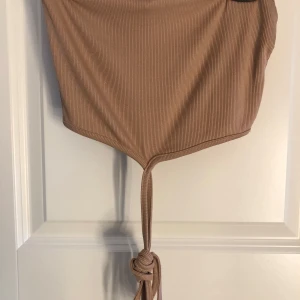 Beige-brun ribbad croppad knyttopp - Säljer en beige-brun ribbad croppad topp från Amisu med knyt framtill och bakom nacken. Toppen har en snygg och trendig design med smickrande passform och långa knytband som kan stylas på olika sätt i storlek s 🤎