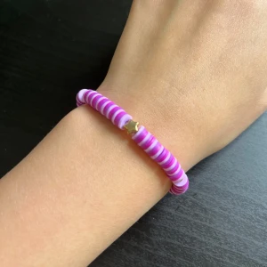 Lila armband med stjärndetalj 💜 - Säljer ett färgglatt armband med platta pärlor i rosa och vitt samt en liten guldfärgad stjärna i mitten. Perfekt accessoar för att liva upp din outfit!💜