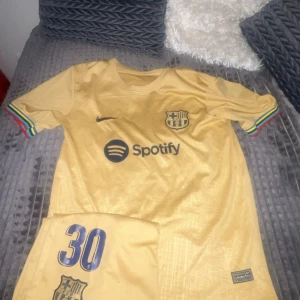 FC Barcelona bortatröja Gavi #30 - Säljer en guldig FC Barcelona bortatröja med Gavi och nummer 30 på ryggen. Tröjan har korta ärmar med färgglada muddar och klubbmärke samt sponsorlogga på bröstet. Perfekt för dig som älskar Barca och vill sticka ut på läktaren! Hör av er vid funderingar😁