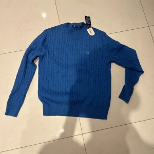 Blå kabelstickad tröja från GANT - Säljer en blå kabelstickad tröja från GANT i modellen Cotton Cable Crew. Tröjan har rund halsringning, ribbade muddar och ett litet broderat GANT-emblem på bröstet. Perfekt för dig som gillar klassisk och stilren design.