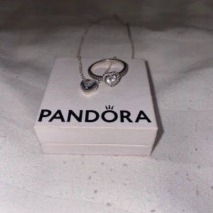 Halsband och ring med hjärta från Pandora - Säljer ett matchande set från Pandora med halsband och ring i silver. Båda smyckena har ett gnistrande hjärta med små stenar runt om. Perfekt för dig som gillar eleganta och romantiska detaljer.