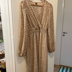 Beige mönstrad långklänning från SHEIN - Beige långklänning från SHEIN med vitt prickigt mönster. Klänningen har lång ärm, v-ringad omlott upptill och resår i midjan för en smickrande passform. Perfekt för dig som gillar en luftig och stilren look.