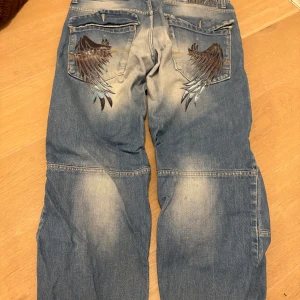 jeans med vingdetalj - Unika blå jeansbyxor från Subsp med stora vingar i svart och blått på bakfickorna. Byxorna har baggy passform, flera fickor och slitna detaljer. Snyggt blekta partier och coola sömmar ger extra edge. Storlek M