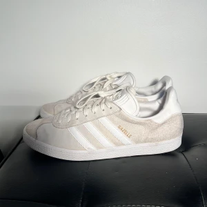 Adidas Gazelle sneakers - Säljer ett par stilrena Adidas Gazelle sneakers i creme/beige färg med klassiska tre ränder på sidan och guldfärgad logga. Skorna har ovandel i mocka och syntet, snörning och platt sula. Perfekta för dig som gillar en clean och tidlös look. Det står på skorna storlek 45 1/3 men dem passar 43-44, jag har storlek 45 och dem är för små för mig. Dem är lite trånga i tårna. 