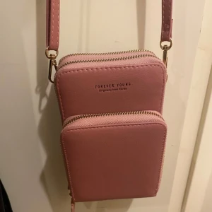 Rosa axelväska från Forever Young - Säljer en söt rosa axelväska från Forever Young med två stora fack och dragkedjor. Väskan har justerbar axelrem och flera kortfack inuti. Perfekt för mobilen och småsaker. Enkel och stilren design med guldiga detaljer.