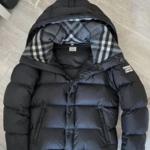 Svart dunjacka från Burberry med rutigt foder - Säljer en svart dunjacka/väst från Burberry med klassiskt rutigt foder i huvan. Jackan har dragkedja och knappar framtill samt två fickor med dragkedja. Perfekt för kyliga och även varma sommar dagar då man kan ta av armarna på den lätt med bara 2 dragkedjor, och har en stilren design med Burberry-detaljer på ärmen.