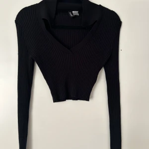 Svart croppad ribbad tröja med v-ringning - Säljer en svart croppad tröja från Bershka med djup v-ringning och långa ärmar. Tröjan är ribbad och sitter tight, perfekt att styla med höga jeans eller kjol. Enkel men snygg design som passar till många outfits.