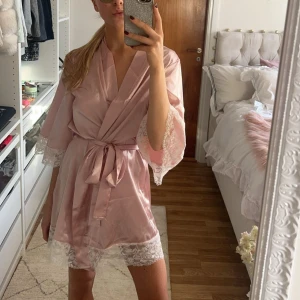 Rosa satin morgonrock med spetsdetaljer - Säljer en söt rosa morgonrock i satin med knytband i midjan och vackra spetsdetaljer vid ärmslut och nederkant. Perfekt för mysiga morgnar eller när du vill känna dig extra lyxig hemma. Köpt second hand så vet tyvärr inte märket