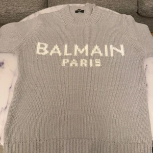 Grå stickad tröja från Balmai Paris - Säljer en grå stickad tröja från Balmai Paris med vit logotyp framtill. Tröjan har rund halsringning och långa ärmar. Perfekt för dig som gillar exklusiva och stilrena plagg.