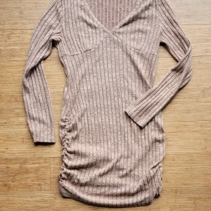 Ribbad långärmad klänning  - Säljer en varmbeige melerad ribbad kort klänning med lång ärm och v-ringad hals. Klänningen har åtsittande passform och snygga rynkade detaljer på sidan. Perfekt för dig som gillar en enkel men stilren look. Använd 1 gång, för liten för mig som har 40-42, uppskattar denna till 38! 