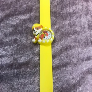 Gul barnklocka med Paw Patrol-motiv - Rolig barnklocka med slap-on-armband i gult och figur från Paw Patrol ovanför urtavlan. Urtavlan har färgglada siffror och motiv av karaktärer från serien. Perfekt för barn som gillar Paw Patrol! 3 år + 