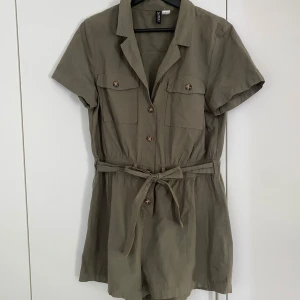 Grön playsuit med knappar och bälte från H&M - Kort grön paysuit från H&M med krage, korta ärmar och knappar framtill. Playsuiten har två bröstfickor och ett matchande knytbälte i midjan för en snygg siluett. .