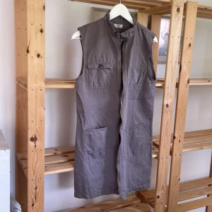 Grå klänning/lång väst från Mads Nørgaard - Säljer en grå/brun klänning eller lång väst från Mads Nørgaard med hög krage och dragkedja framtill. Västen har fyra fickor både upptill och nedtill 